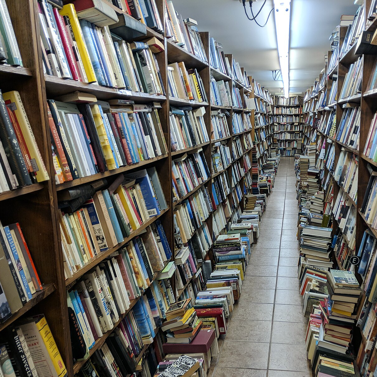 A narrow aisle lined with densely packed shelves and stacks of used books in a secondhand bookshop.