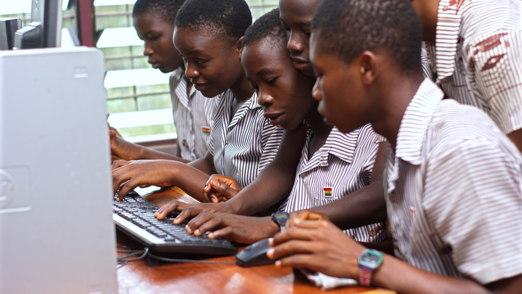Six students in school uniforms gather around a desktop computer in a classroom.